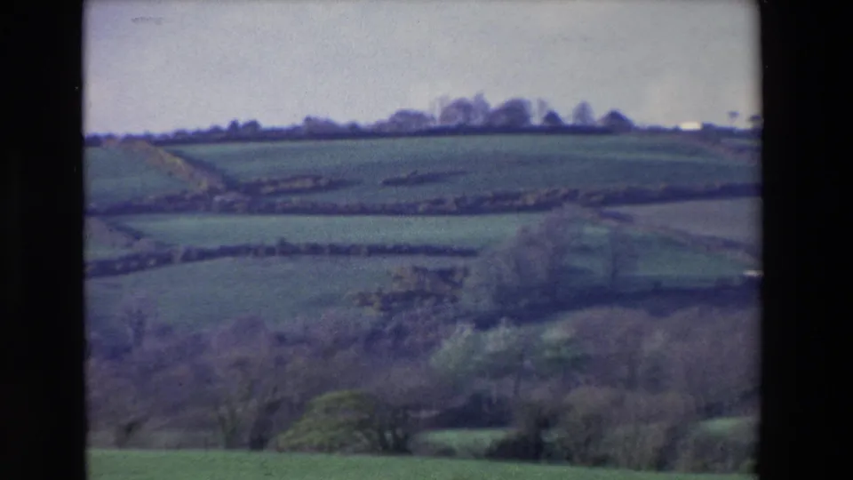 1969 home-movie frame from Ireland showing green hillside fields, hedgerows, foreground woodland, and a small pale structure near the horizon.