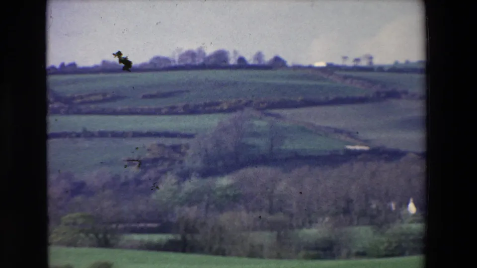 A 1969 8mm countryside view in Ireland with divided fields, scattered trees, distant pale structures, and visible film specks across the frame.
