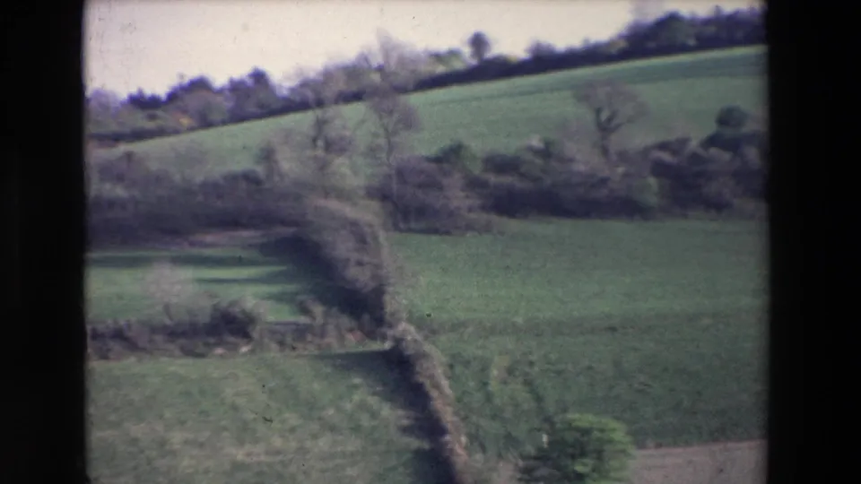 1969 Ireland 8mm frame showing a tilted view across green fields, hedgerows, bare trees, and layered field boundaries in soft home-movie focus.