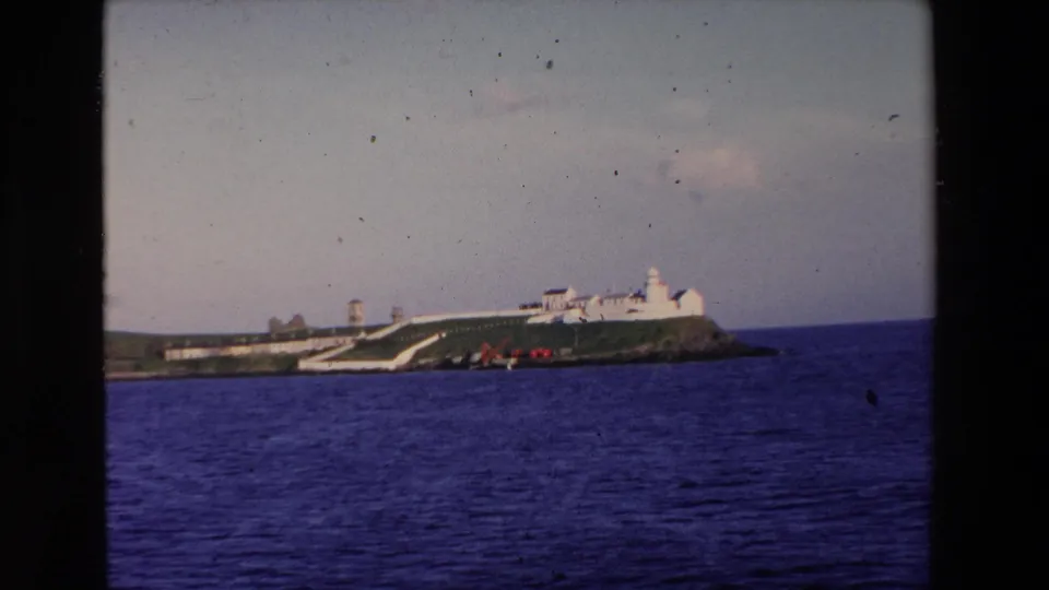 Coastal Ireland view from 1969 showing a low island with white buildings and a lighthouse-like tower across blue sea.