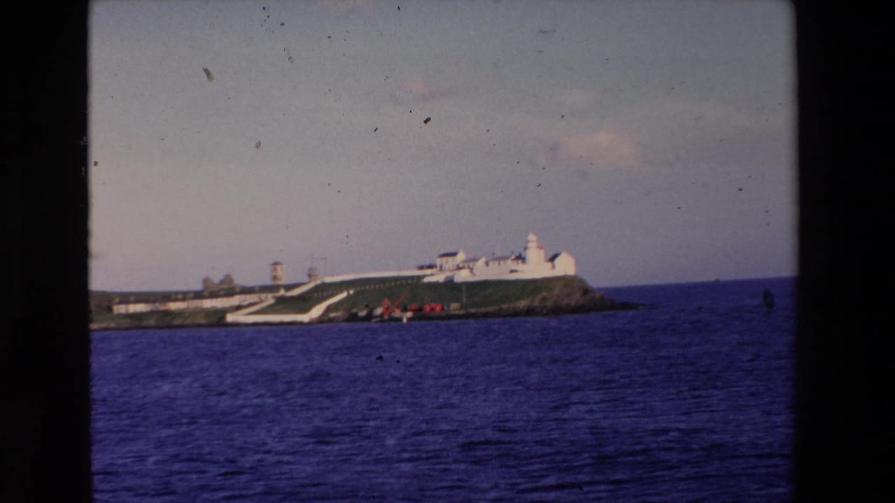 1969 Ireland 8mm frame showing a distant white lighthouse complex and buildings on a grassy rocky island across deep blue ocean.
