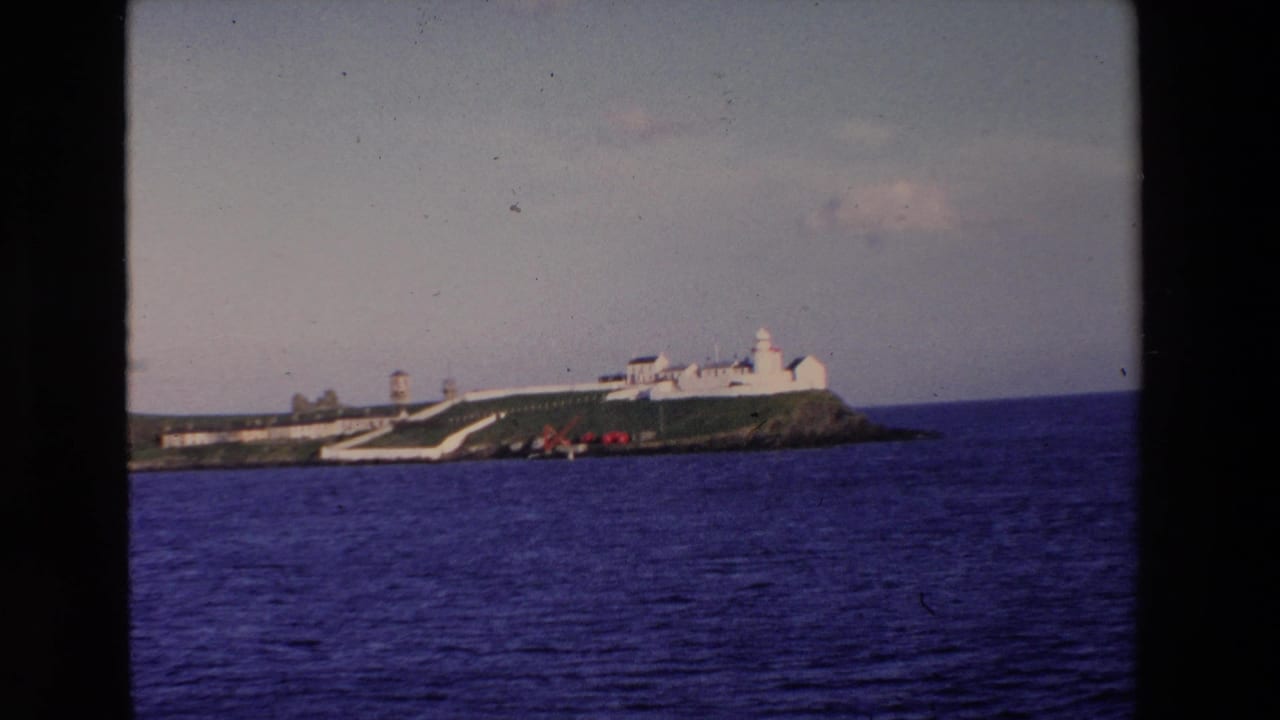 1969 Ireland home-movie frame with a white lighthouse complex on a low rocky island, seen across blue water under a pale sky.
