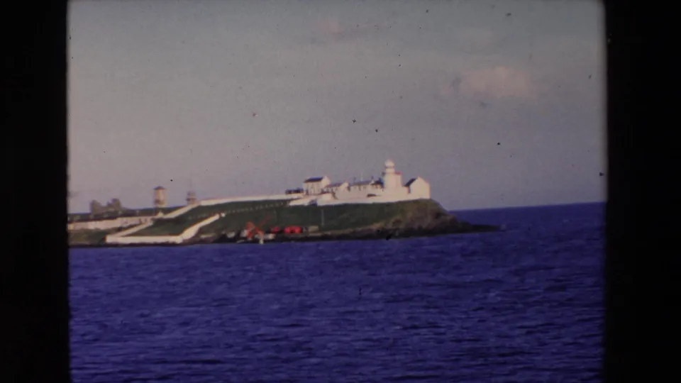 1969 Ireland 8mm frame showing the lighthouse complex from a slightly closer or shifted angle, with ocean foreground and small red objects along the rocky edge.