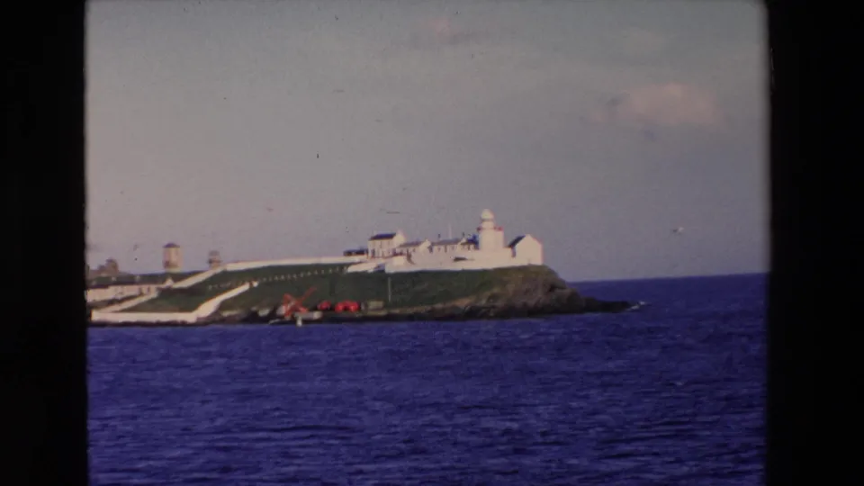 1969 Ireland home-movie frame of a white lighthouse complex on a grassy rocky island, with blue ocean in the foreground and soft sky behind it.