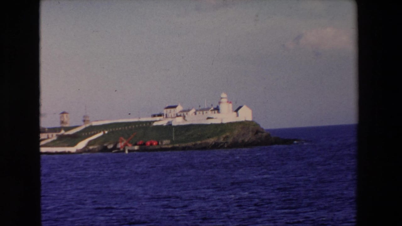 1969 Ireland 8mm frame showing a distant white lighthouse complex on a grassy rocky island with blue water in the foreground.