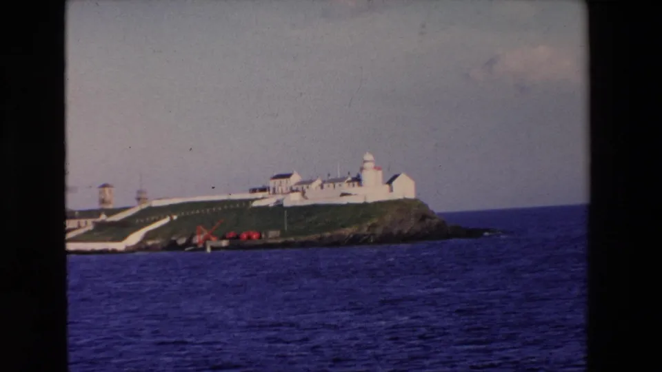 1969 Ireland 8mm frame showing a distant white lighthouse complex on a grassy rocky island with blue water in the foreground.