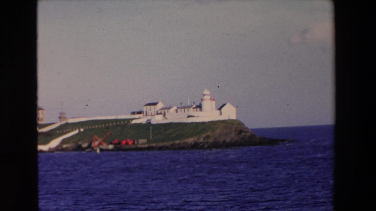 1969 Ireland 8mm frame with a white lighthouse station and connected buildings on a green island, seen from across deep blue water.