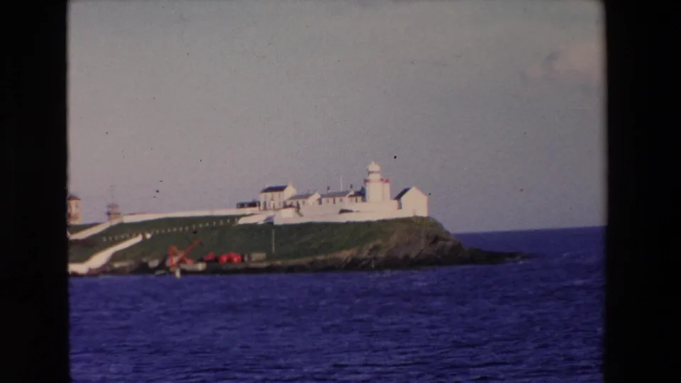 1969 Ireland 8mm frame with a white lighthouse station and connected buildings on a green island, seen from across deep blue water.