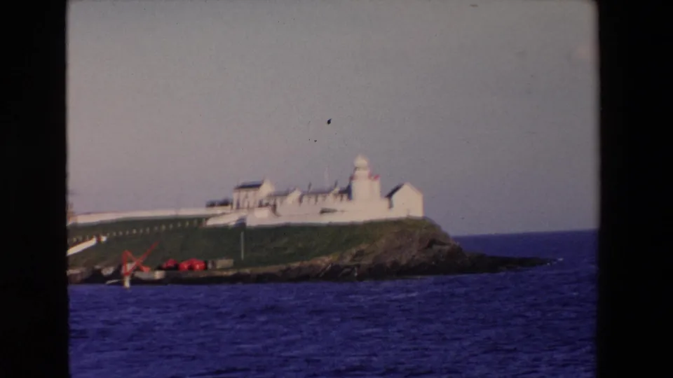 1969 Ireland 8mm frame showing the lighthouse complex partly cropped by the film edge, with grassy terrain, rocks, and ocean below.