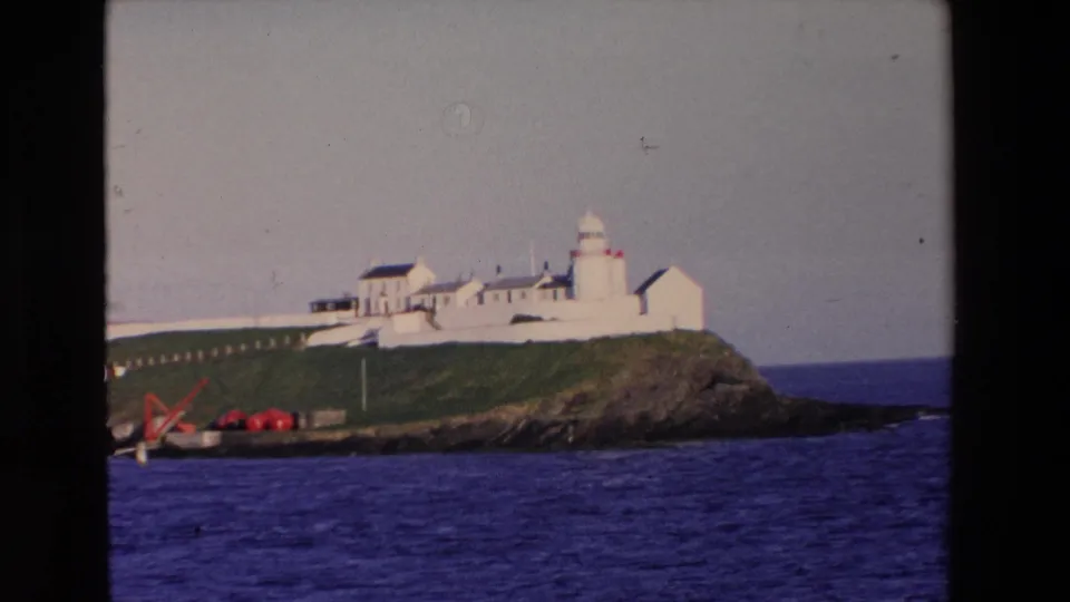 1969 Ireland 8mm frame showing a white lighthouse complex on a grassy rocky island with red equipment near the shore and blue ocean in front.