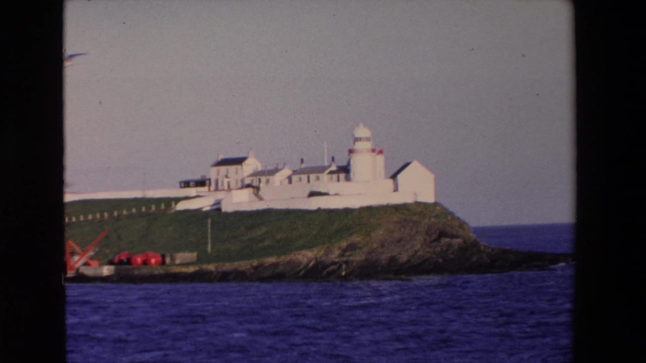 1969 8mm frame from Ireland showing a white lighthouse complex and whitewashed buildings on a green rocky headland overlooking blue water.