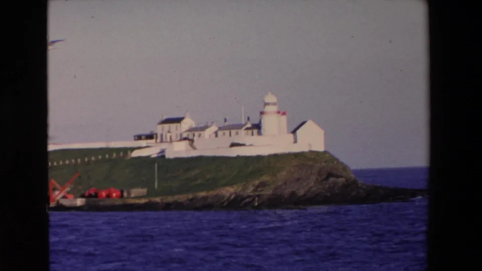1969 8mm frame from Ireland showing a white lighthouse complex and whitewashed buildings on a green rocky headland overlooking blue water.