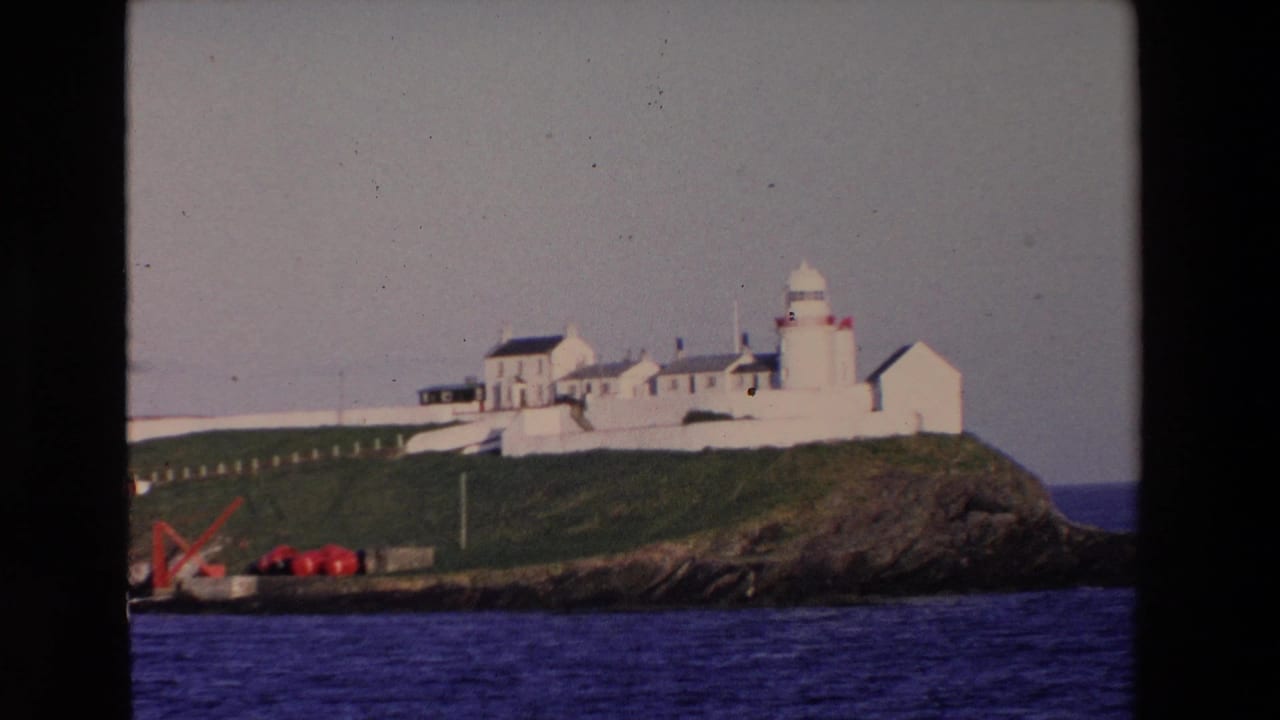 1969 Ireland travel footage frame with a white lighthouse and nearby buildings standing on a grassy, rocky outcropping above deep blue water.