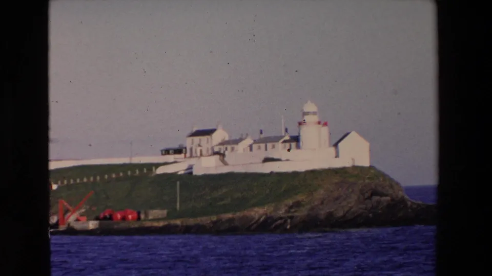 1969 Ireland travel footage frame with a white lighthouse and nearby buildings standing on a grassy, rocky outcropping above deep blue water.