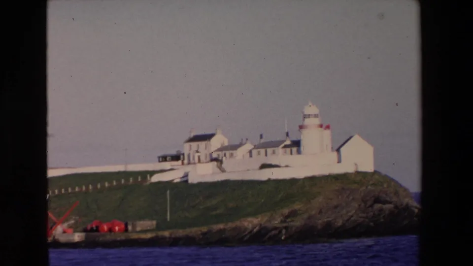 A slightly closer 1969 8mm view of an Irish lighthouse complex, with white buildings on a grassy headland and blue ocean below.