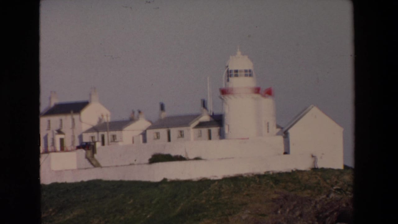 1969 Ireland 8mm frame showing a white lighthouse with red accents beside whitewashed buildings and a perimeter wall on a grassy coastal headland.