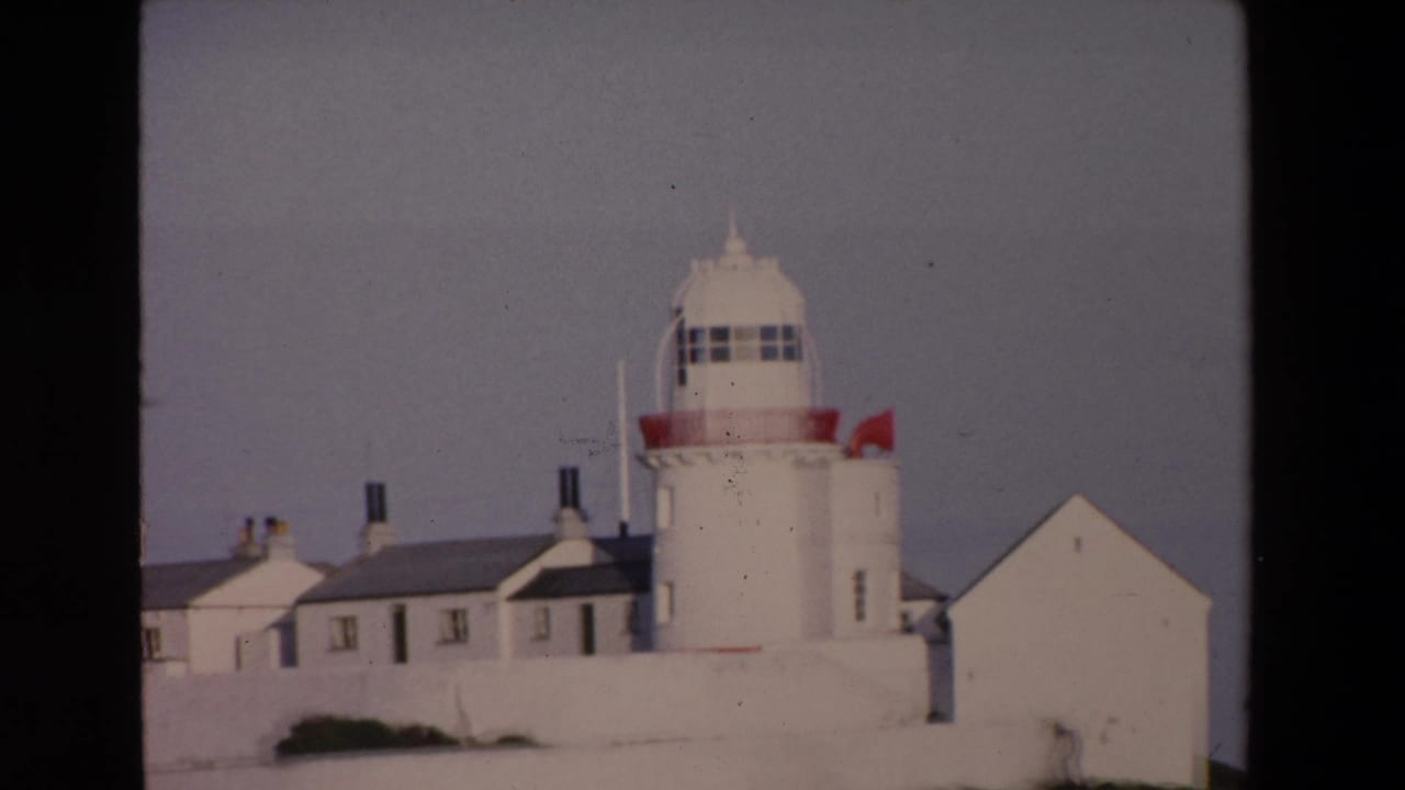 White lighthouse tower with adjoining white buildings and a wall beneath a pale sky.