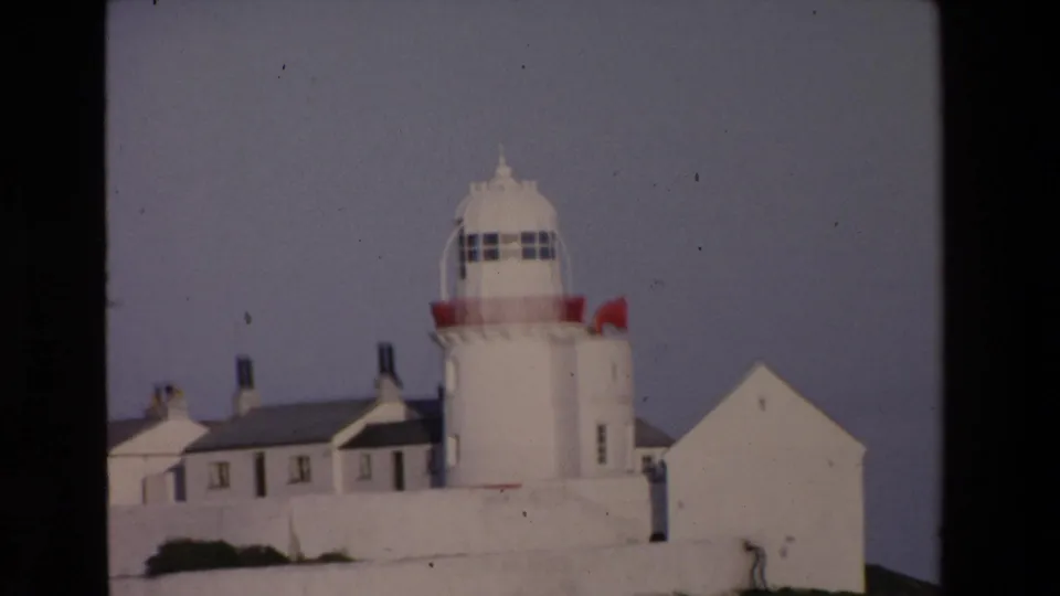 1969 Ireland 8mm frame showing a closer visitor view of the lighthouse complex, with the tower centered above whitewashed buildings and a gabled outbuilding.