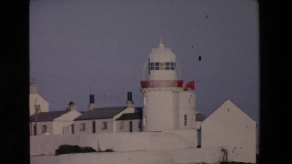 1969 Ireland home-movie frame of a white coastal lighthouse complex with red accents, adjoining white buildings, chimneys, wall, and soft 8mm grain.