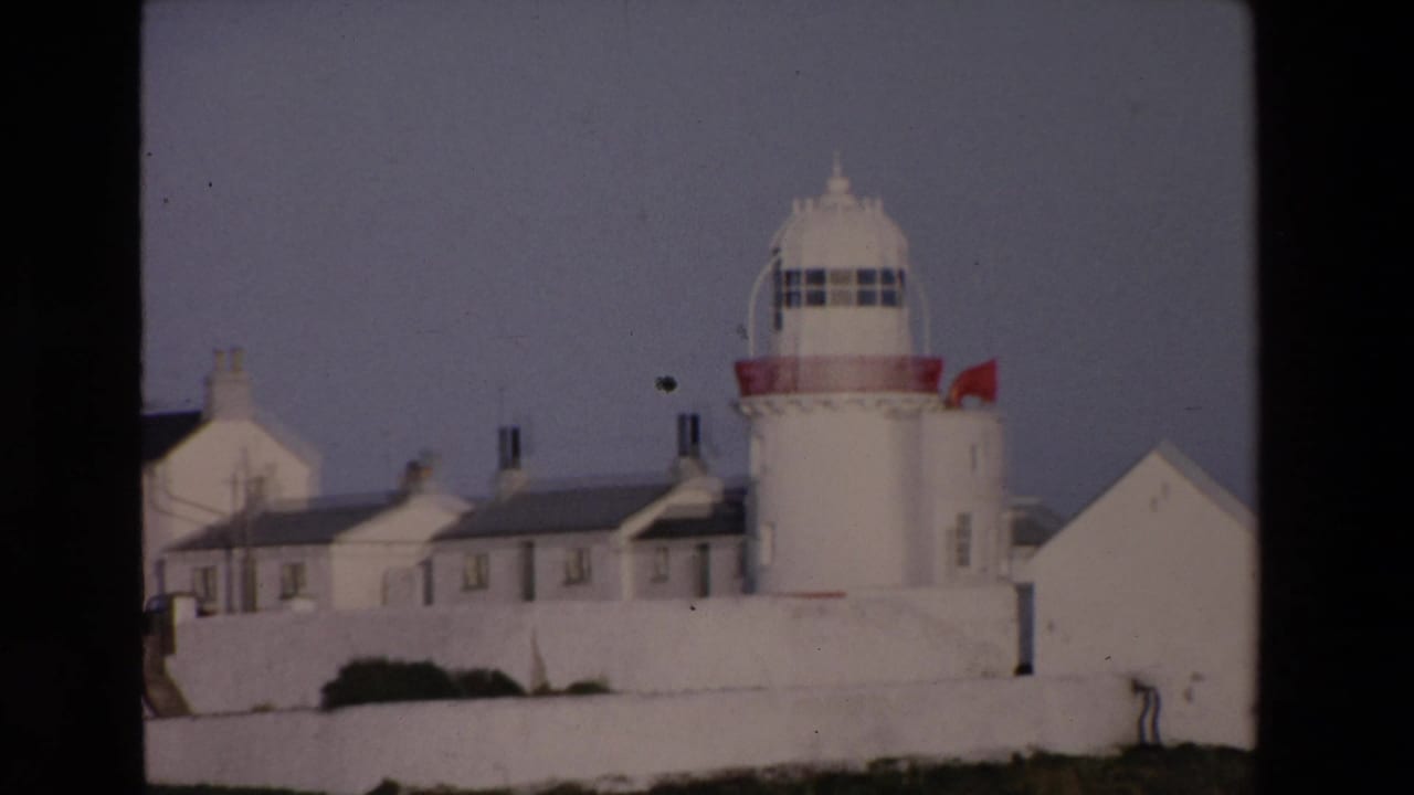 1969 Ireland 8mm frame showing a white lighthouse with a red band, adjoining white buildings, dark roofs, and a small flag under a subdued sky.