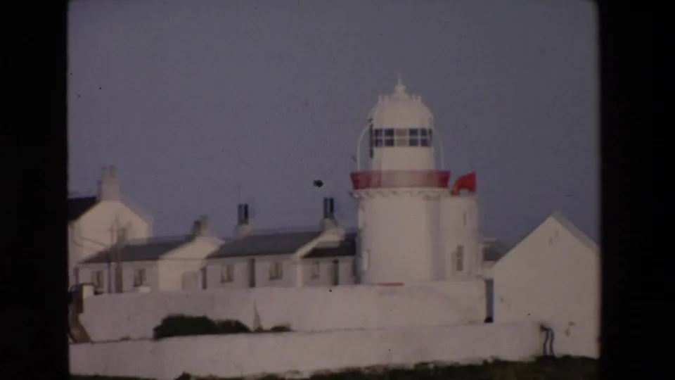 1969 Ireland 8mm frame showing a white lighthouse with a red band, adjoining white buildings, dark roofs, and a small flag under a subdued sky.