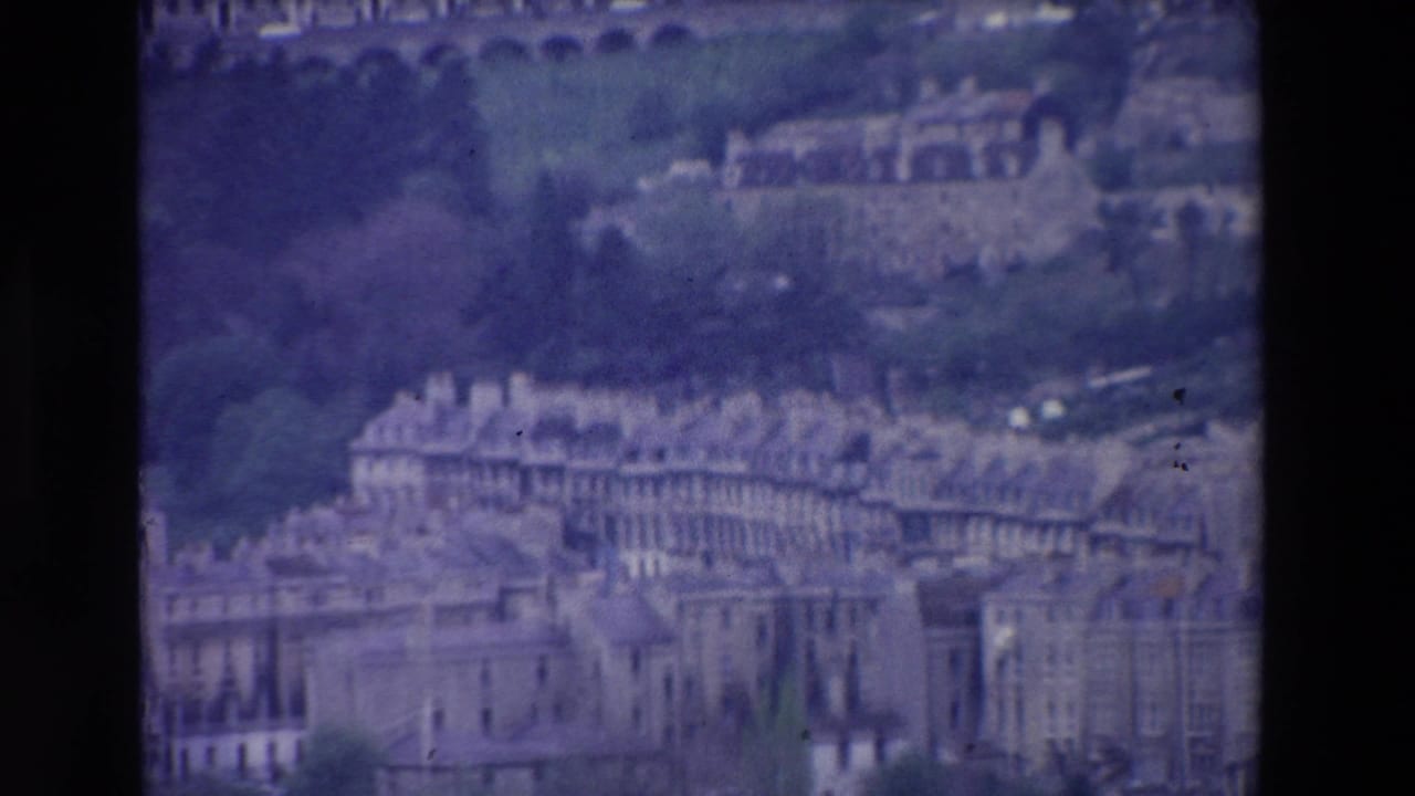 Grainy 1969 Ireland home-movie frame showing a hillside town view with rows of older buildings, rooflines, greenery, and distant arched structures.