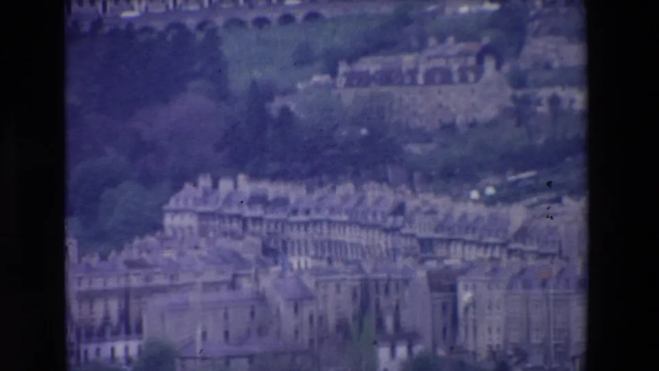 Grainy 1969 Ireland home-movie frame showing a hillside town view with rows of older buildings, rooflines, greenery, and distant arched structures.