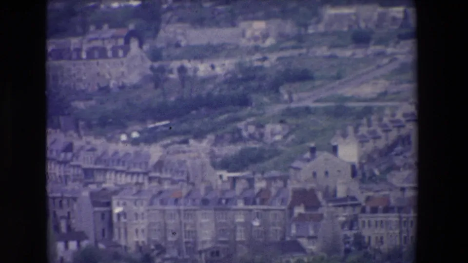 Grainy 1969 Ireland home-movie still showing rooftops in the foreground, a green hillside behind them, and larger older structures across the upper frame.