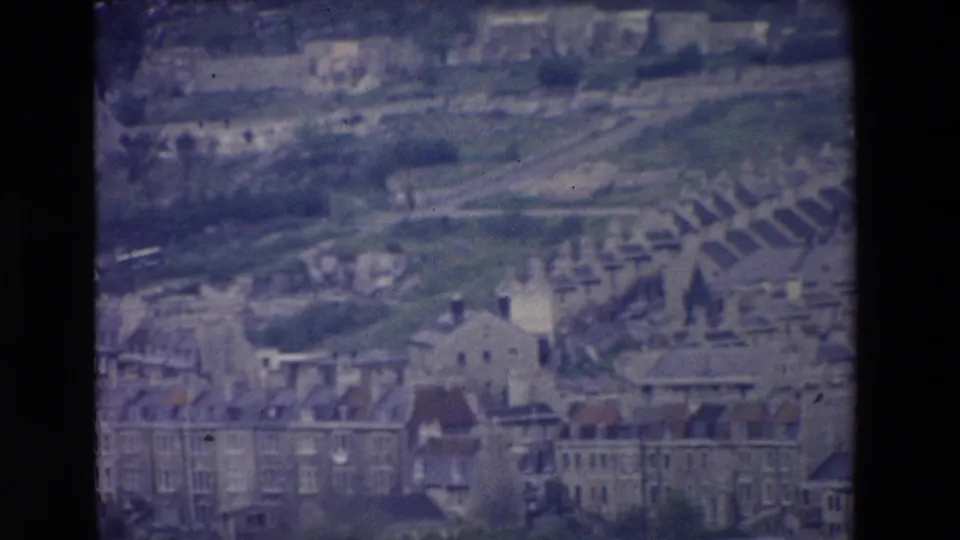 1969 Ireland 8mm frame showing an elevated panoramic view of older town buildings, layered rooflines, green slopes, and distant arched forms under a faded color cast.