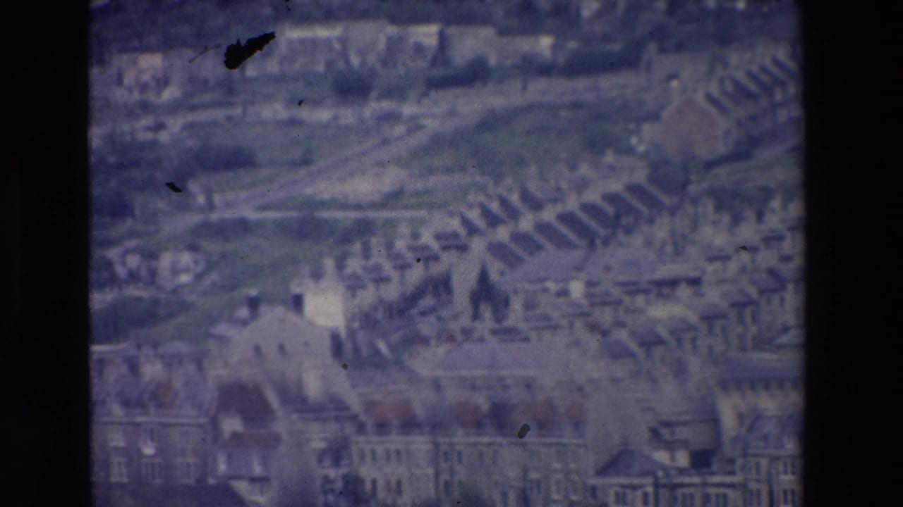 1969 Ireland 8mm frame showing a distant hillside town, dense rows of houses, larger buildings, and a church spire under a muted sky.