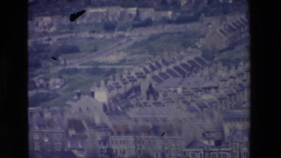 1969 Ireland 8mm frame showing a distant hillside town, dense rows of houses, larger buildings, and a church spire under a muted sky.