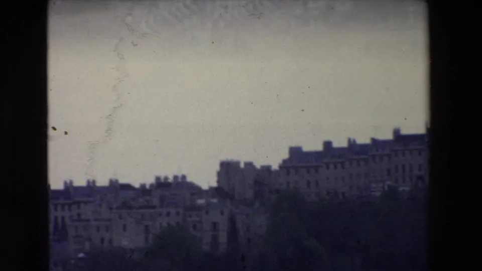 1969 Ireland 8mm frame showing a lower city skyline with tree cover in the foreground and long masonry buildings silhouetted against a pale sky.