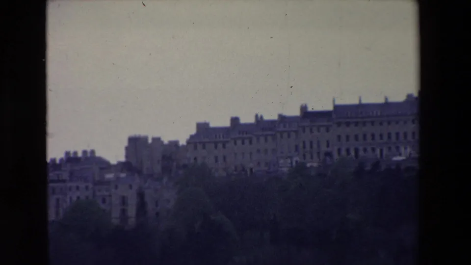 1969 Ireland home-movie frame showing a wider town skyline, dark trees in the foreground, clustered buildings, and a small bird in the pale sky.