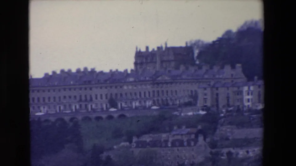 1969 Ireland home-movie frame showing stone townhouses on a slope, a larger building above them, trees, parked cars, and smaller structures in the lower foreground.