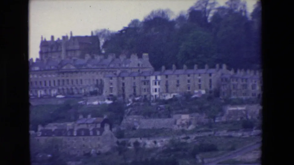 1969 Ireland home-movie frame showing a hillside townscape with stone buildings, wooded upper ground, smaller structures below, and a road or path crossing the foreground.