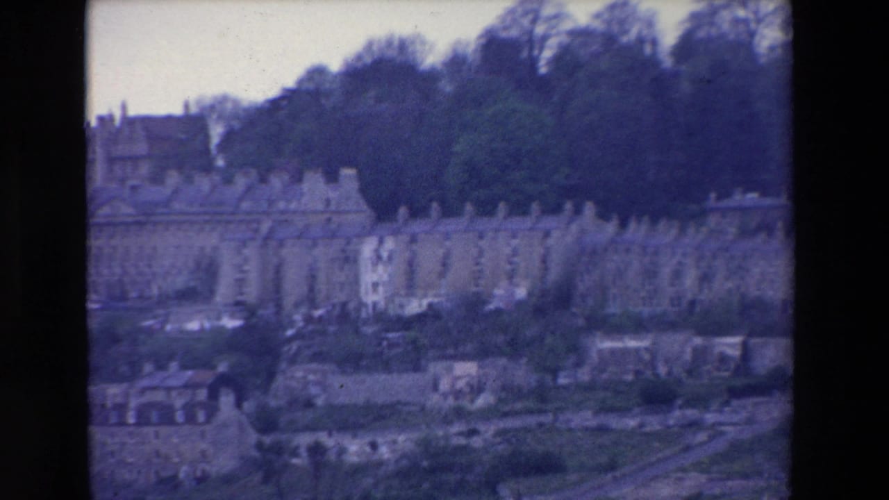 Distant 1969 Ireland 8mm view of light-colored terraced buildings, roofs, open ground, and tall trees on a hillside.