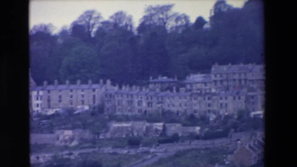 1969 Ireland home-movie frame showing a hillside neighbourhood with terraced houses, larger multi-storey buildings, trees, and a road at the lower edge.