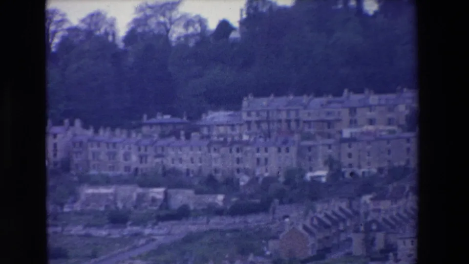 Soft 8mm frame from Ireland in 1969 showing residential blocks, terraced houses, dark trees, and foreground rooftops during a slow pan.
