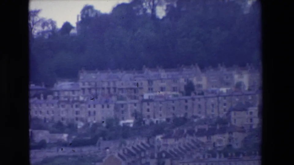 Final 1969 Ireland frame with a distant, blue-purple 8mm view of multi-storey buildings, rooflines, trees, and a hillside skyline.