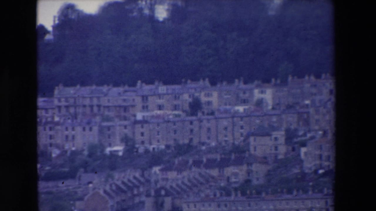 1969 Ireland 8mm frame showing pale terraced buildings spread across a hillside beneath dense trees, with a blue-purple film cast and dark frame edges.