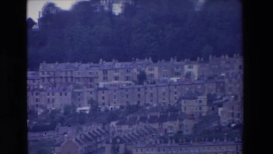 1969 Ireland 8mm frame showing pale terraced buildings spread across a hillside beneath dense trees, with a blue-purple film cast and dark frame edges.