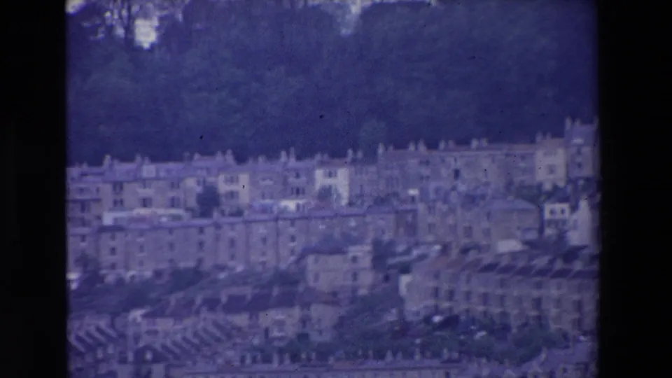 Elevated 1969 Ireland home-movie view of closely packed terraced buildings and rooftops beneath a wooded ridge, softened by grain and color shift.