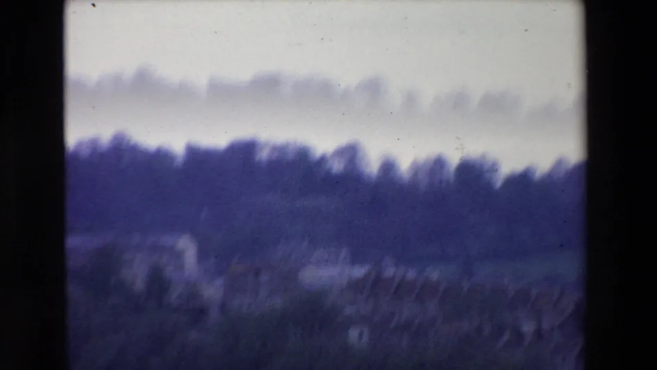 Soft, motion-blurred 8mm frame from the same Irish hillside view, with dark tree silhouettes, grassy slope, pale sky, and indistinct buildings below.