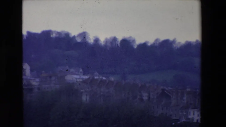 1969 Ireland 8mm frame showing a distant hillside townscape with scattered pale buildings, dark trees along the ridge, open grass, and soft home-movie exposure.