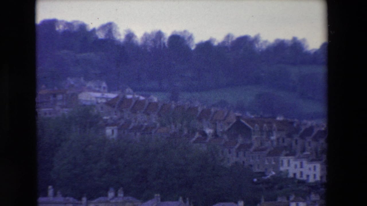 1969 Ireland 8mm frame showing a distant row of houses beneath green fields and a dark tree line, with soft focus and blue film cast.