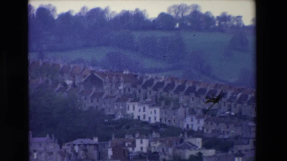 1969 Ireland 8mm view of clustered hillside houses below open green fields, with soft focus, visible grain, and a small film blemish near the lower center.
