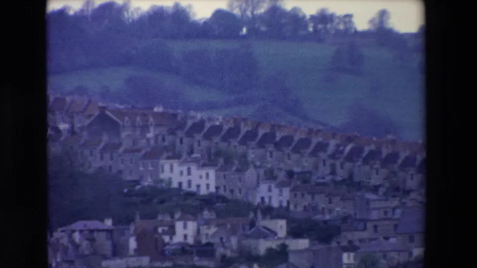 1969 Ireland home-movie frame showing hillside houses and rooftops beneath green fields and a dark tree line, with blue-cast 8mm softness.