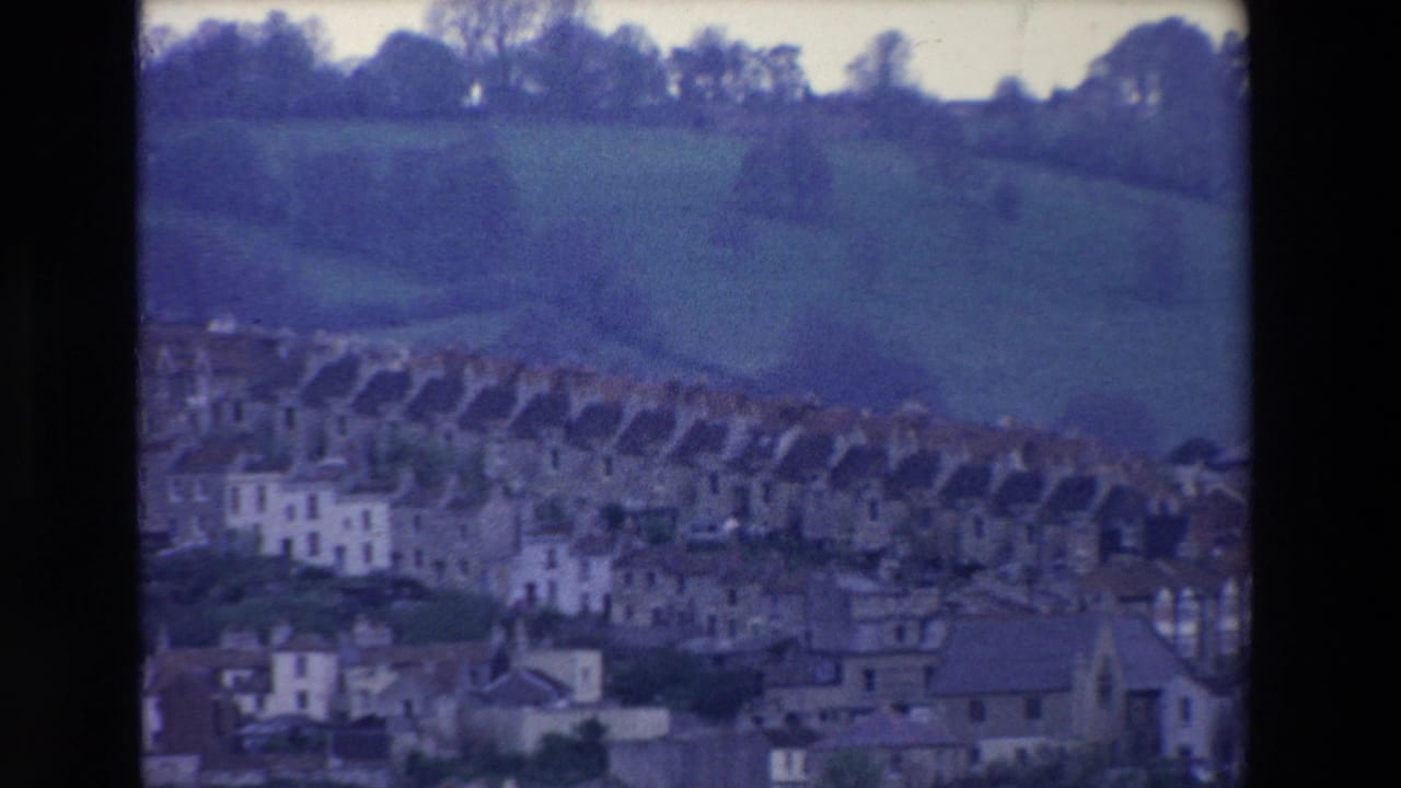 1969 Ireland 8mm frame showing rows of pale houses clustered below green hillside fields, seen from a distant visitor-shot viewpoint.