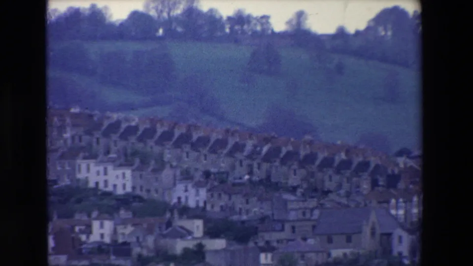 1969 Ireland 8mm frame showing rows of pale houses clustered below green hillside fields, seen from a distant visitor-shot viewpoint.