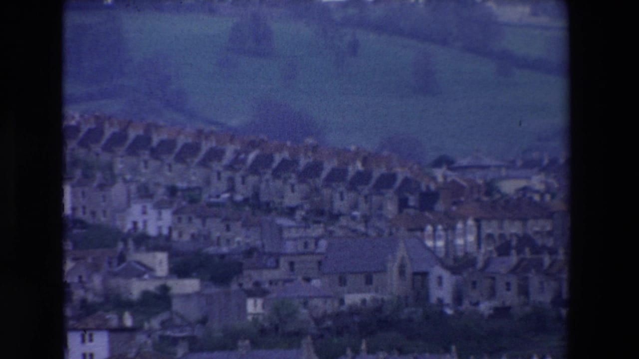 Distant 8mm view of an Irish hillside settlement in 1969, with repeated rooftops, pale building fronts, and fields rising behind the houses.